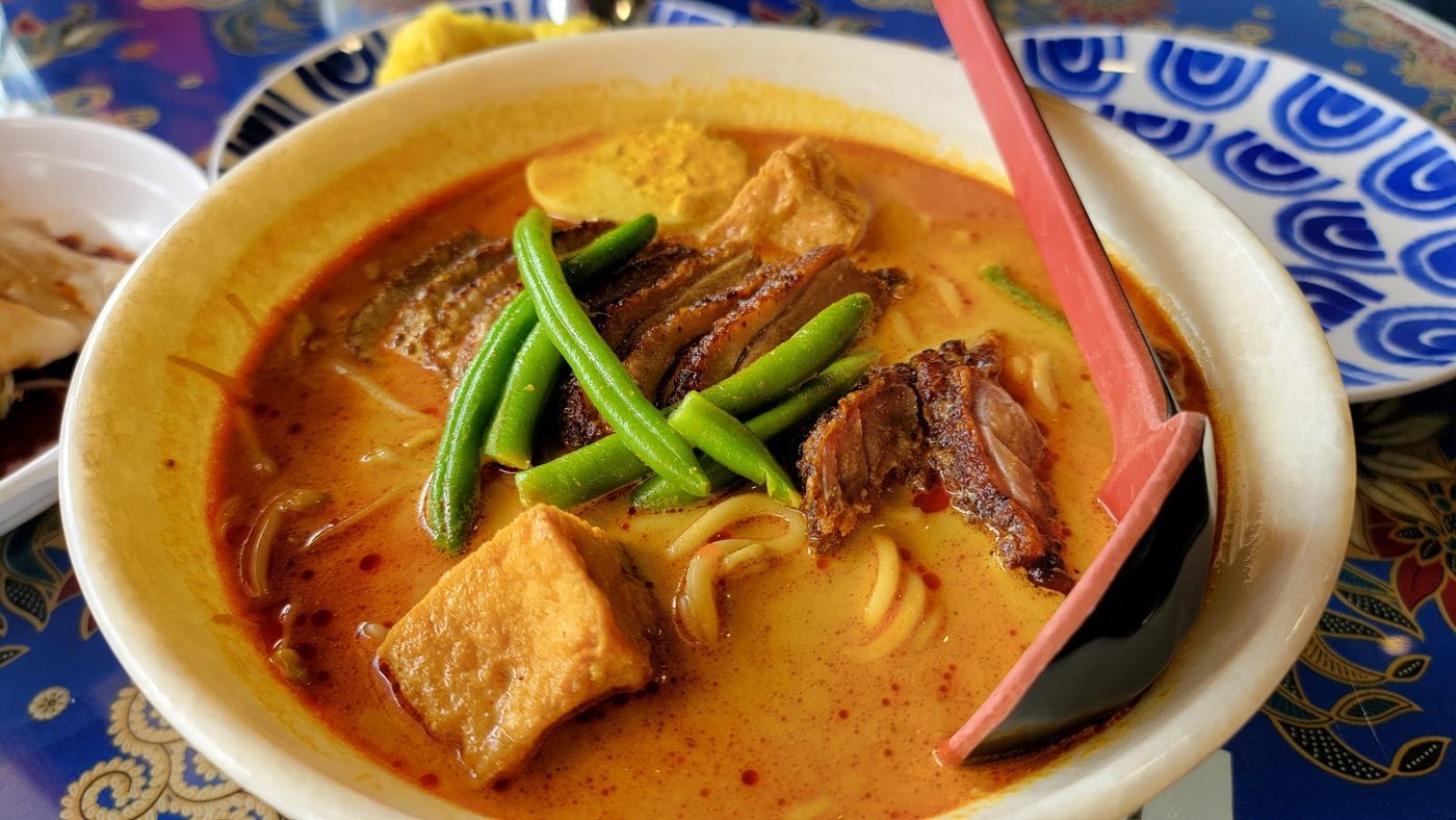 A deep bowl of noodles in red laksa broth with vegetables and toppings
