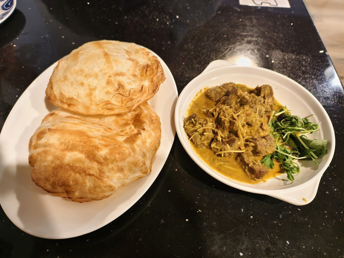 Two pieces of roti canai beside a bowl of curry and greens